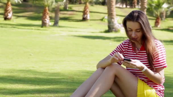 Hermosa mujer sentada en la hierba en el parque de la ciudad de verano con teléfono
 - Imágenes, Vídeo