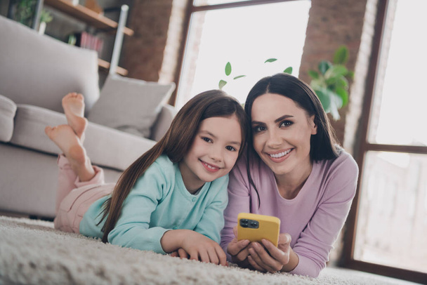 Photo of sweet pretty daughter mom dressed casual clothes lying floor texting modern gadget smiling indoors apartment room. - Foto, afbeelding