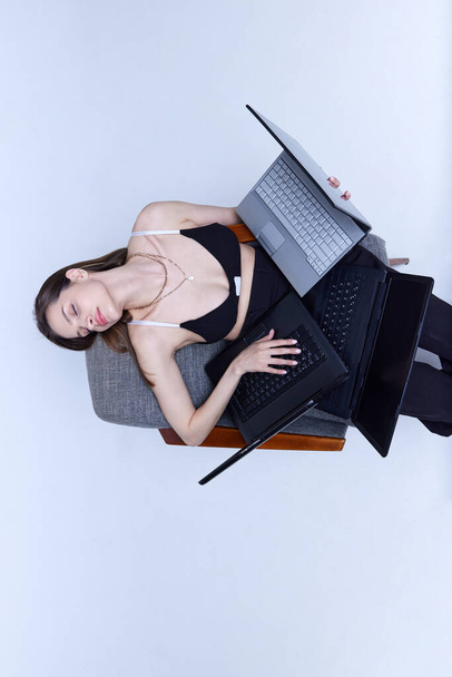 Young girl sitting on chair with many laptops and working against studio background. Freelancer, creator. Concept of fashion, 80s, 90s style, retro and vintage, gadgets, business, technology - Foto, imagen