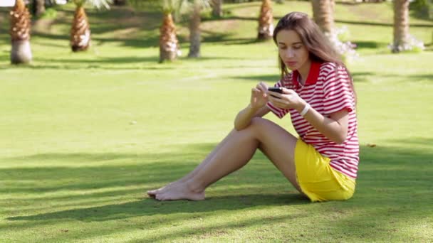 Beautiful woman sitting on grass in summer city park with phone - Filmati, video