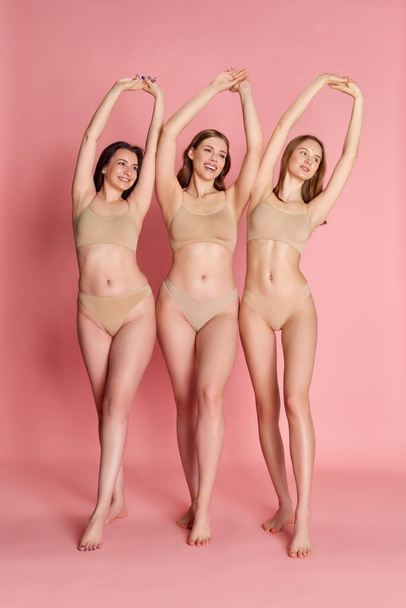 Νεαρό σώμα. Πορτρέτο με ομάδα χαρούμενων χαμογελαστών κοριτσιών, γυναίκες που φορούν μπεζ εσωτερική φθορά ποζάρουν πάνω από ροζ φόντο στούντιο. Έννοια της φροντίδας του δέρματος, υγιεινή διατροφή, πλαστική χειρουργική, καλλυντικά, διαφήμιση - Φωτογραφία, εικόνα