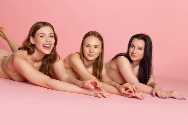 Portrét se skupinou tří okouzlujících dívek, ženy v béžovém vnitřním oblečení ležící na růžovém pozadí. Zavřít. Koncepce péče o tělo a pokožku, zdravé stravování, plastická chirurgie, kosmetika, reklama - Fotografie, Obrázek