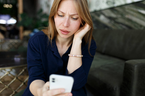 Young woman sad, reading bad news email, break up message from ex-boyfriend on smartphone sitting lonely on couch in living room. Depressed unhappy female desperate of heartbreak sms on mobile phone. - Photo, Image