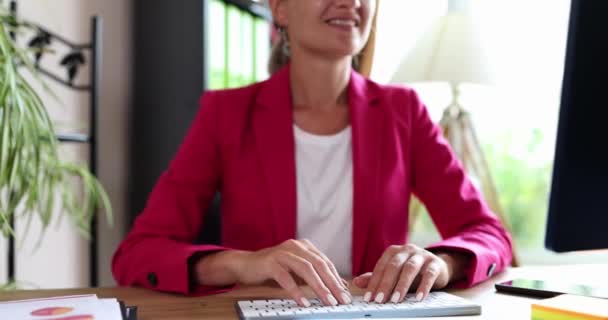 Manager vrouw in roze jas zit aan kantoor tafel voor de computer en glimlacht. Affable werknemer golven hand om nieuwe klant slow motion begroeten - Video