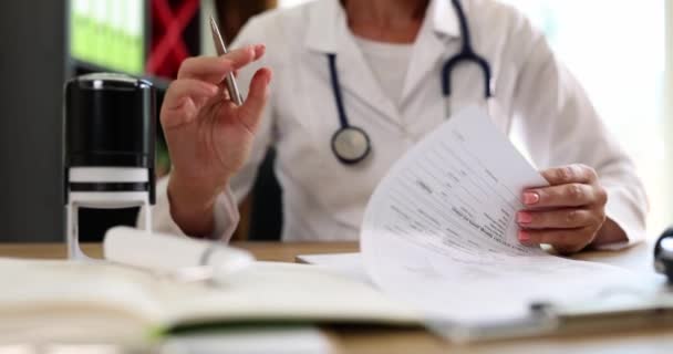 Vrouwelijke arts zet stempel op papieren met de diagnose met behulp van inkt afdrukken. Vrouw met stethoscoop werkt zittend aan tafel in ziekenhuis kantoor slow motion - Video