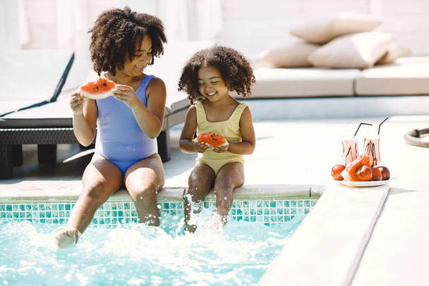 Two curly multiracial sisters sitting near the pool. Girls wearing blue and yellow swimwear. Girls splashing the water in pool and holding a peace of watermelon. - Foto, immagini
