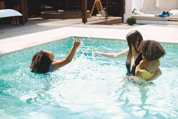 Two curly multiracial sisters and their caucasian mother playing in the pool. Girls wearing blue and yellow swimmengwear and mother wearing a black one. Woman and girls splashing the water. - Photo, Image