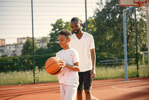 Young father and his son playing basketball in basketball court near the park. Man and boy wearing white t-shirts. Boys holding an orange basketball ball with black stripes.l - Fotografie, Obrázek