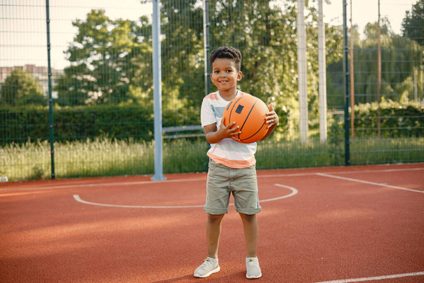 Young boy standing on basketball court near the park. Boy wearing white t-shirt. Boy playing with basketball ball. - Foto, Imagen
