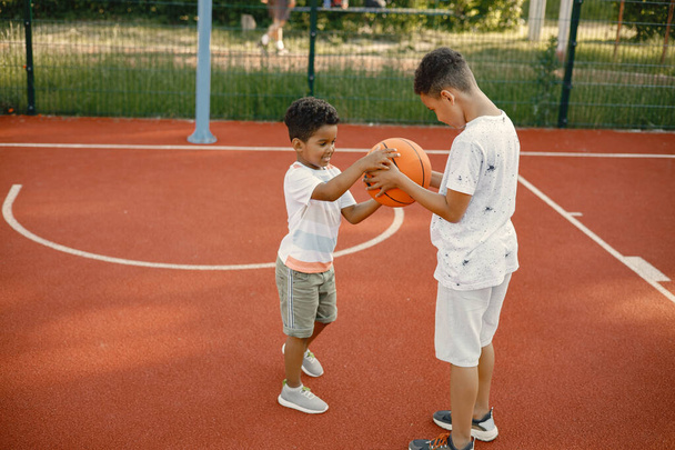 Two multiracional brothers playing basketball on a court near the park. Boys wearing white t-shirts. Older brother teach little one how to play basketball. - Φωτογραφία, εικόνα