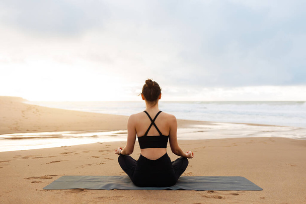Poranna medytacja. Nierozpoznawalna spokojna kobieta w stroju aktywnym ćwicząca jogę na świeżym powietrzu, pani siedząca w pozycji lotosu na macie fitness na plaży w pobliżu oceanu, ciesząca się medytacją - Zdjęcie, obraz