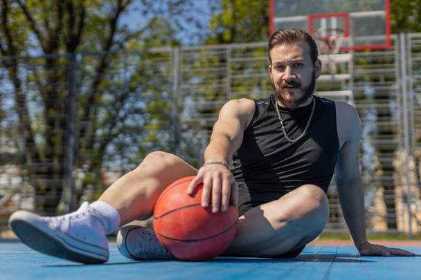 バスケットボールゲームをプレイした後、スポーツウェアの疲れ運動性レバノン人男性の肖像画。都会の公園裁判所でリラックスした保持ボールに座っている男。スポーツ分野でのフィットネスルーチン。やる気だ。屋外 - 写真・画像