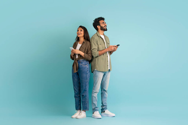 Randi mobil alkalmazás koncepció. Stílusos fiatal keleti férfi és nő hipszterek állnak háttal, okostelefonok színes stúdió háttér, csevegés online, nézi a másolás tér, teljes hossz - Fotó, kép