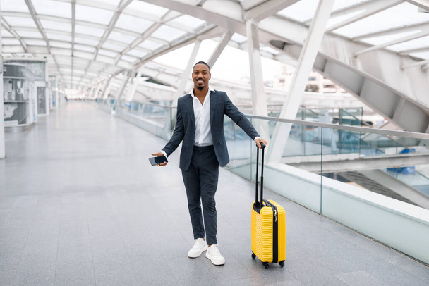 Hombre negro sonriente en elegante traje de pie con maleta en la terminal del aeropuerto, Guapo joven afroamericano posando en la cámara mientras espera la profundidad del vuelo, sosteniendo el teléfono inteligente en la mano - Foto, imagen