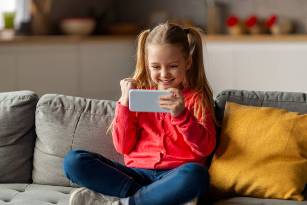 Nettes kleines Mädchen, das zu Hause Spiele auf dem Smartphone spielt und Erfolge feiert, fröhliche Preteen-Mädchen, die die Faust schütteln und Ja sagen, Online-Gewinn genießen, während sie auf der Couch im Wohnzimmer sitzen - Foto, Bild