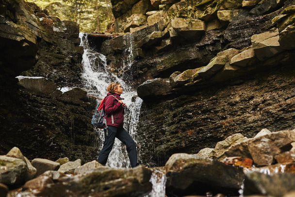 Hiking in mountains. Woman enjoying hike on sunny vacation day. Female with backpack walking close to waterfall. Spending summer vacation close to nature - Fotografie, Obrázek