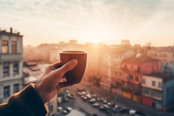 Mies, jolla on kuppi kuumaa kahvia tai teetä käsissään näkymä hämärtynyt kaupunkikuva. Mies katolla tai parvekkeella kuumalla juomalla aamulla auringonvalossa ja kaupunkikuva taustalla. Generatiivinen tekoäly - Valokuva, kuva