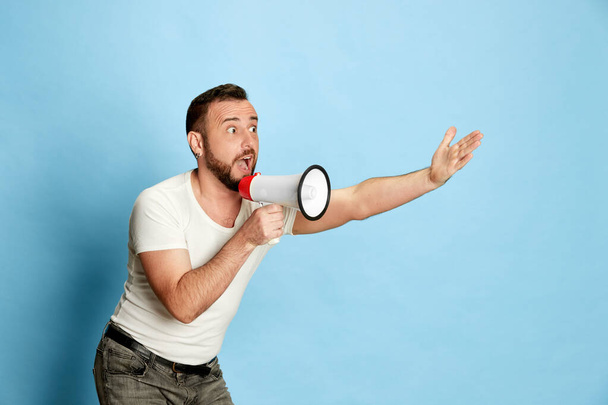 Portrait of emotional bearded man in casual white t-shirt shouting in megaphone against blue studio background. Concept of human emotions, lifestyle, facial expression, news, sport fan, betting. Ad - Photo, Image