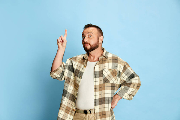 Portrait of bearded man in casual clothes, checkered shirt raising finger up, expressing ideas against blue studio background. Concept of human emotions, lifestyle, facial expression. Ad - Foto, Bild