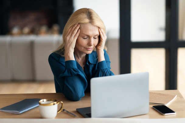 Sad mature lady sitting at table in front of computer, suffering from headache, menopause, stress in living room interior. Problems in work, business, deadline with device at home - Foto, Imagen