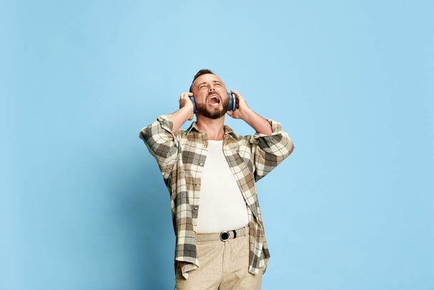 Portrait of bearded man in white t-shirt and checkered shirt listening to music in headphones and singing against blue studio background. Concept of human emotions, lifestyle, facial expression. Ad - Valokuva, kuva