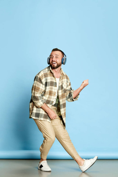 Portrait of bearded man in white t-shirt and checkered shirt listening to music in headphones against blue studio background. Concept of human emotions, lifestyle, facial expression. Ad - Foto, imagen