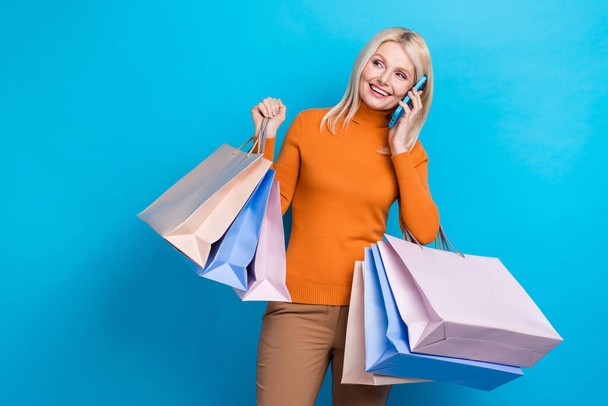 Portrait of charming attractive businesswoman blond hair orange sweatshirt talking on phone hold bags isolated on blue color background. - Fotó, kép