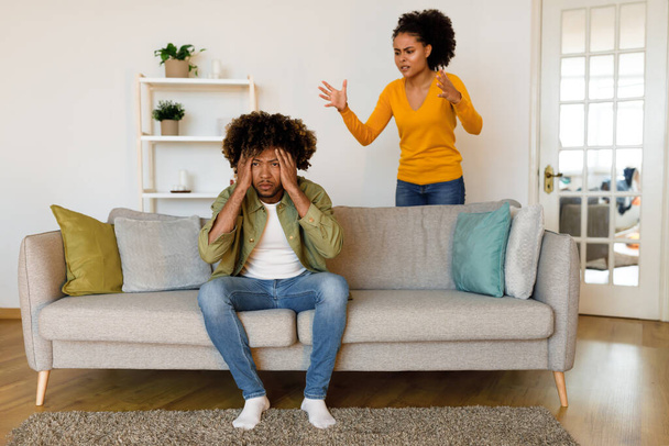 Married Black Couple Having Quarrel At Home. Angry Wife Shouting At Unhappy Husband Having Marital Crisis In Living Room. Man Covering Ears Tired Of Family Conflicts. Selective Focus - Foto, Imagen
