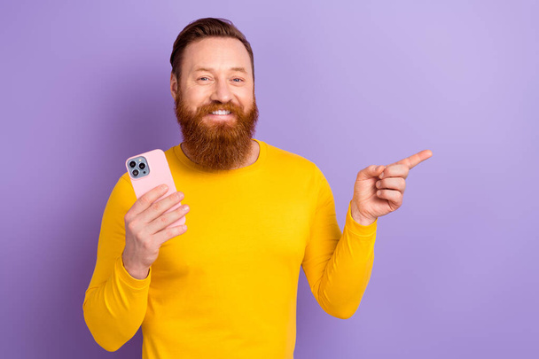 Photo of young blogger guy redhair beard masculine wear yellow pullover hold phone direct finger twitter isolated on violet color background. - Valokuva, kuva