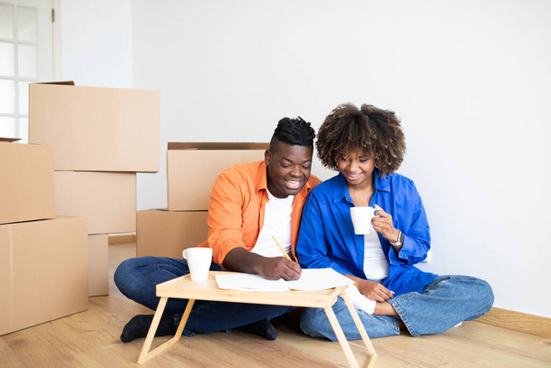 Молодая черная пара планирование бюджета на ремонт дома после переезда, улыбаясь афроамериканских супругов, сидя среди картонных коробок, пить кофе и записи в блокноте, подсчет расходов, копировать пространство - Фото, изображение