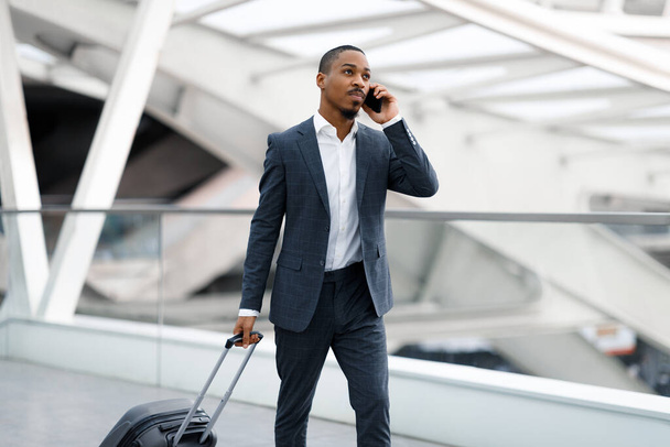 afro-americano empresário falando no celular enquanto caminhava com bagagem no aeroporto, jovem preto masculino no terno ter telefone celular conversa ao ir para a porta de voo, espaço de cópia - Foto, Imagem