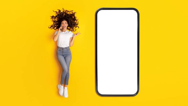Mobilní nabídka. Veselá žena mluví na telefonu ležící poblíž Big Smartphone s prázdnou obrazovkou přes žluté Studio pozadí, Top View Shot. Dáma komunikuje přes mobil. Panorama, Mockup - Fotografie, Obrázek