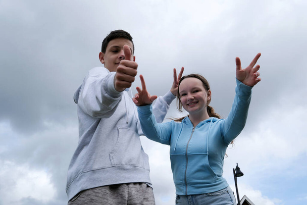 couple of teenagers a boy and a girl stand against the background of a sky with clouds raise their thumbs up build horns to each other spread their fingers laughing having fun having fun approving - Фото, зображення