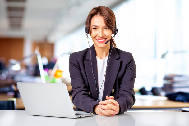 Customer service assistant wearing headset while sitting behind her computer and working in the call center. Attractive business woman wearing black blazer. - Zdjęcie, obraz