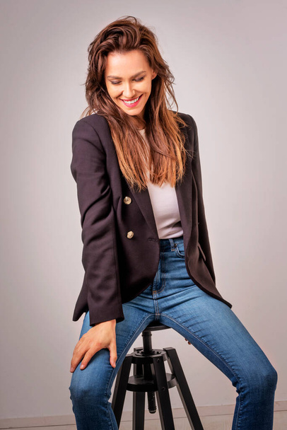 Smiling brunette businesswoman sitting against grey background. Confident female is wearing black blazer and blue jeans. She is having brown hair. Copy space. Studio shot. - Фото, изображение