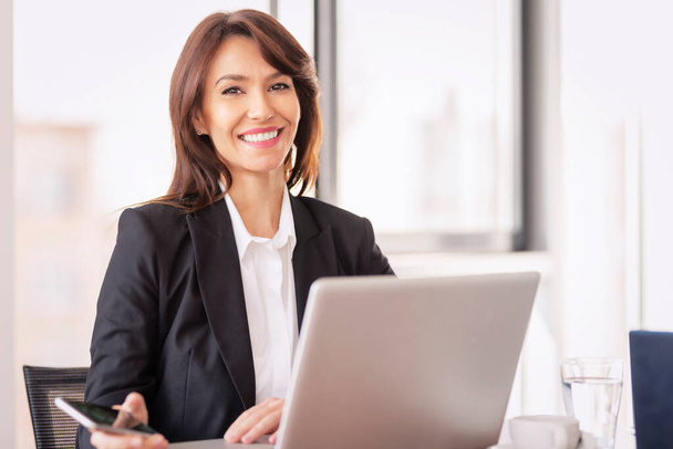 Attractive businesswoman wearing blazer and using laptop for work. Confident female looking at camera and cheerful smiling.  - Foto, Imagen