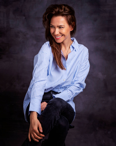 Studio portrait of attractive woman wearing shirt and jeans while sitting at isolated dark background. Mid aged brunette haired female cheerful smiling. Copy space.  - Valokuva, kuva