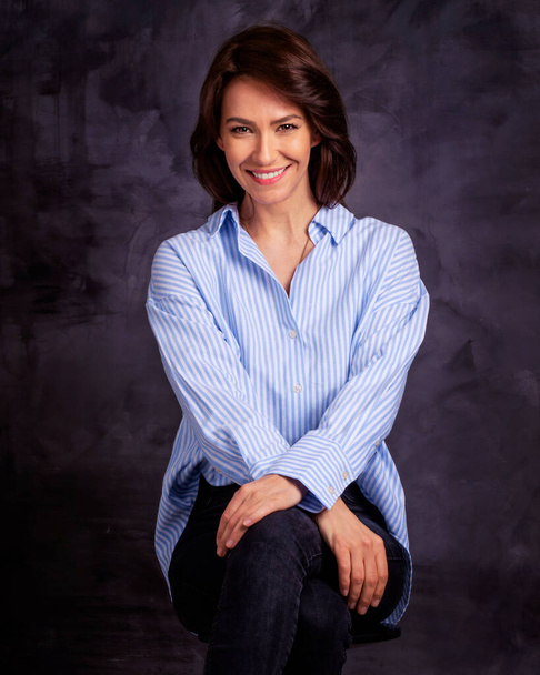 Studio portrait of attractive woman wearing shirt and jeans while sitting at isolated dark background. Mid aged brunette haired female cheerful smiling. Copy space.  - Φωτογραφία, εικόνα