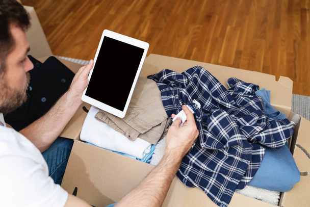 Pantalla en blanco de tableta digital en manos masculinas y caja de cartón con ropa, ropa de inventariado de hombre para organización benéfica.  - Foto, imagen