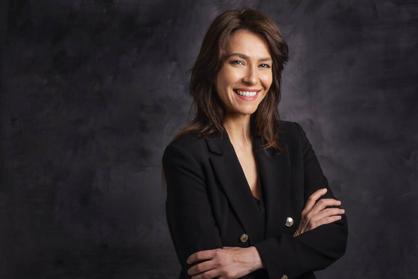 Close-up of a cheerful smiling middle aged woman posing at isolated dark background. Brunette haired female wearing blazer. Copy space. Studio shot. - 写真・画像