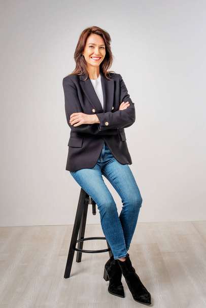 Studio portrait of attractive woman wearing blazer and blue jeans while sitting at isolated background. Full length shot. Copy space.  - Foto, Imagem