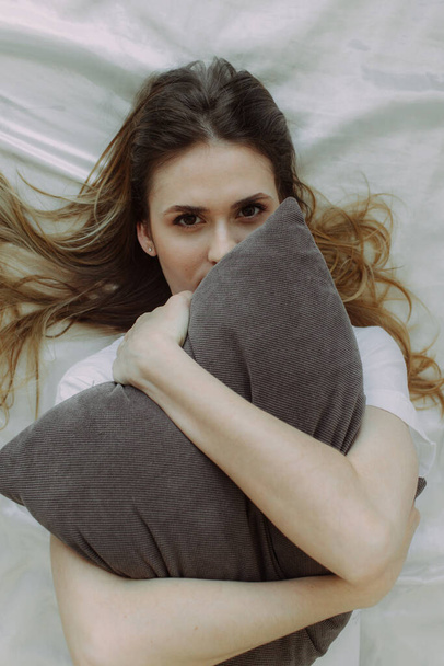 Genç bir kadın rahat bir yatakta ipek çarşaflarla yastığa sarılıyor. Rahatlama ve sağlıklı uyku kavramı. - Fotoğraf, Görsel