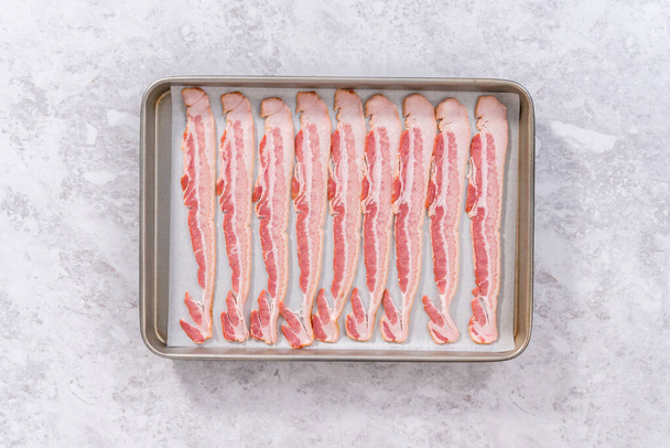 Flat lay. Uncooked bacon strips on a baking sheet with white parchment paper. - Foto, imagen