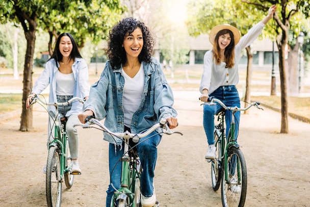Young group of multiracial women having fun riding bike together in the park. Three smiling female friends enjoying time together on summer vacation. Friendship and holidays concept. - Photo, Image