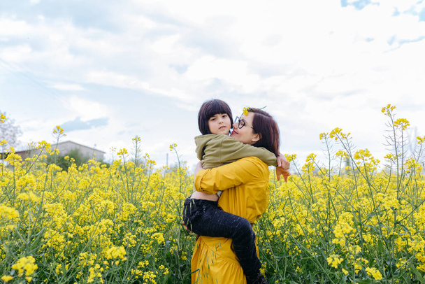 mother in yellow dress holding a child with short dark hair in arms in a field with yellow flowers - Photo, Image