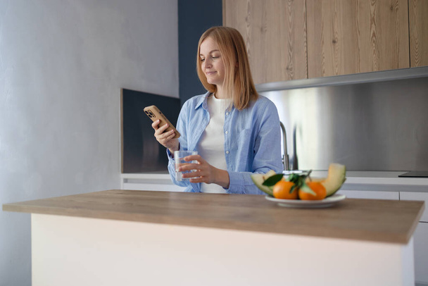 Улыбающаяся женщина в синей стильной рубашке смотрит на свой мобильный телефон и пьет воду, стоя на современной кухне. Утренние привычки и ритуалы. Зависимость от интернета, концепция планирования дня. Высокий - Фото, изображение
