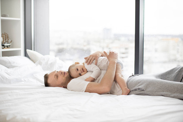 Side view of touching moment of pure love between young father and little daughter lying in embrace on pristine white bed, near panoramic window offering expansive view. Concept of unbreakable bond. - 写真・画像