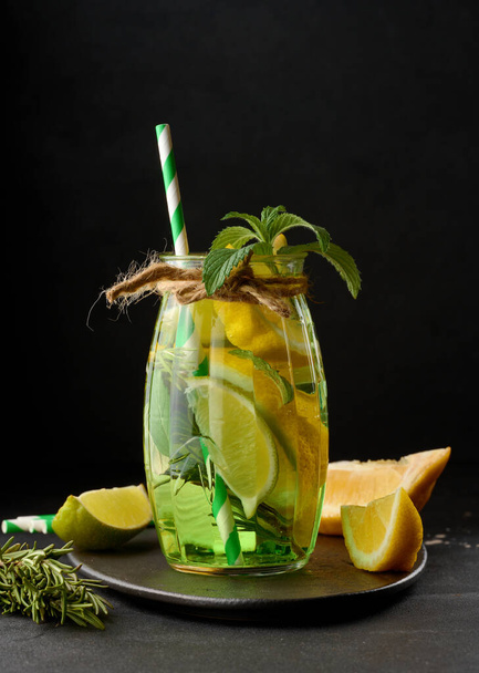 Лимонад в прозрачном стакане с лимоном, лаймом, веточками розмарина и листьями мяты на черном фоне - Фото, изображение