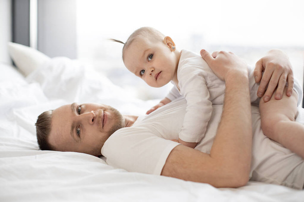 Portrait of handsome bearded man holding little kid on belly. Kind father with adorable baby girl in white clothes lying on soft bedding and looking at camera. Concept of parental love and care. - Valokuva, kuva