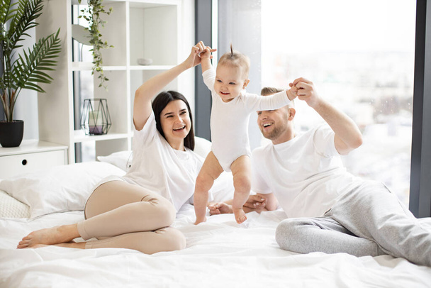 Loving mom and dad holding hands of active girl and helping baby to take first steps on large white bed. Cheerful laughing married couple in casual outfit playing with joyful child. - Foto, afbeelding